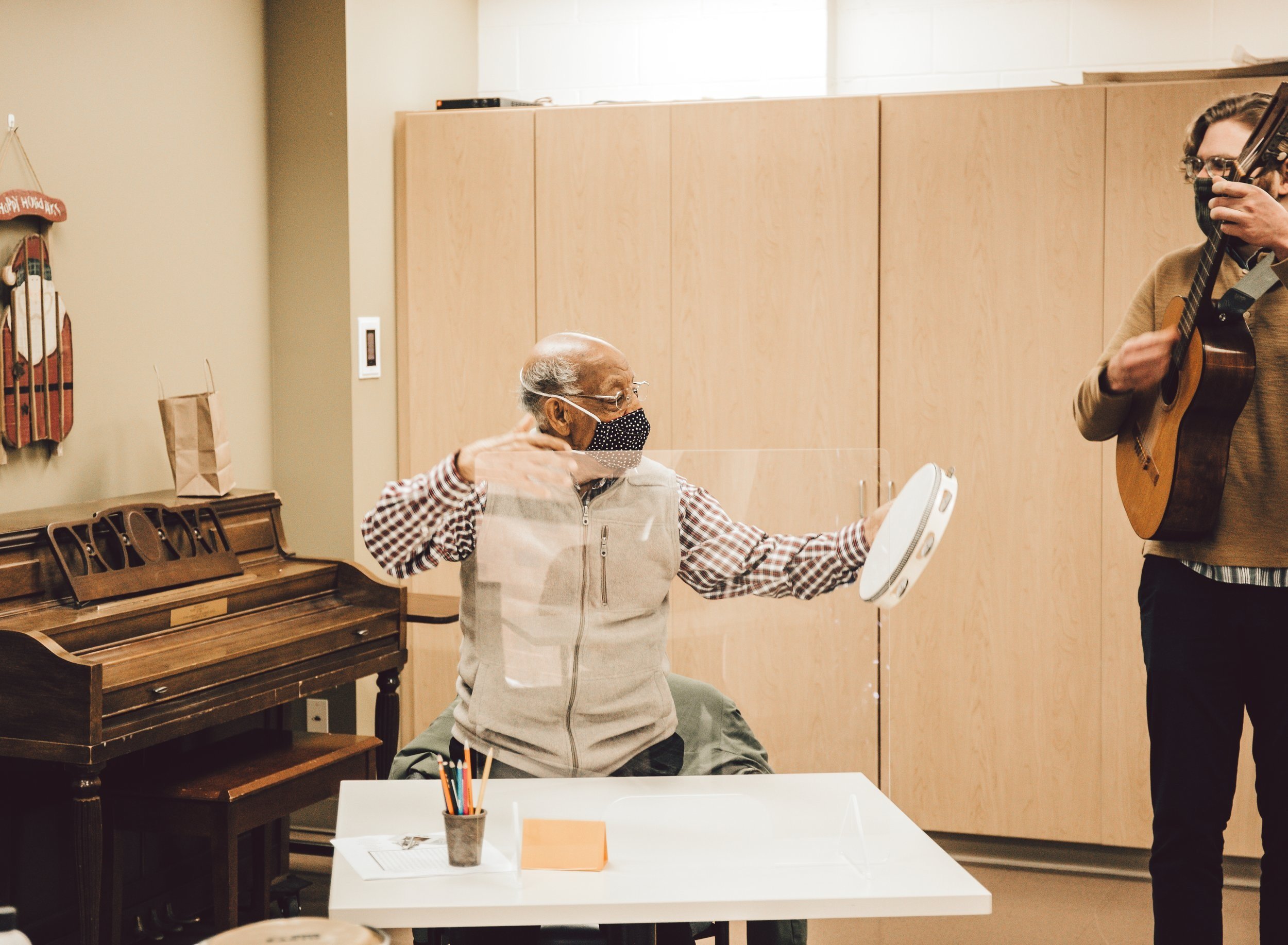 An elderly person in light clothing gesturing expressively while a musician plays guitar in the background